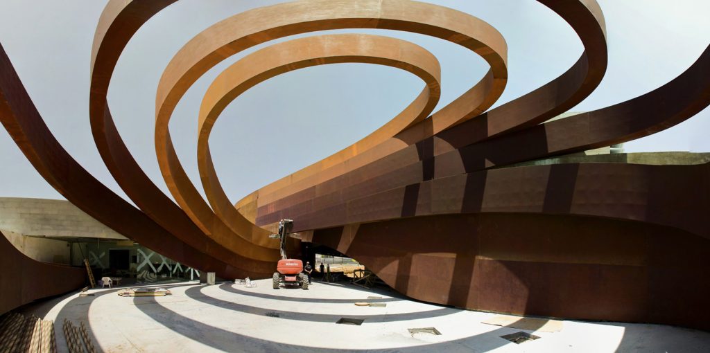 An architectural curved structure rising up roof-like from the courtyard underneath, above a construction vehicle