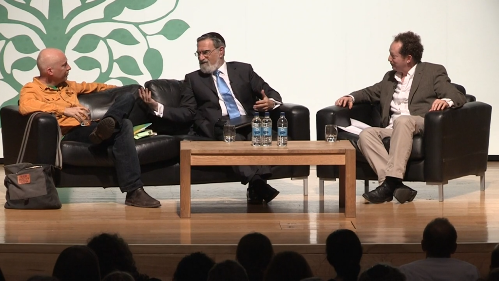 three people speak to each on chairs in front of a live audience