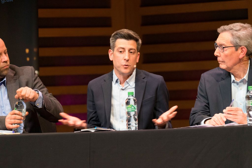 Three men in dark suits seated at a table with a table cloth and microphones. The central figure is gesturing with open hands and has a bottle of water in front of him