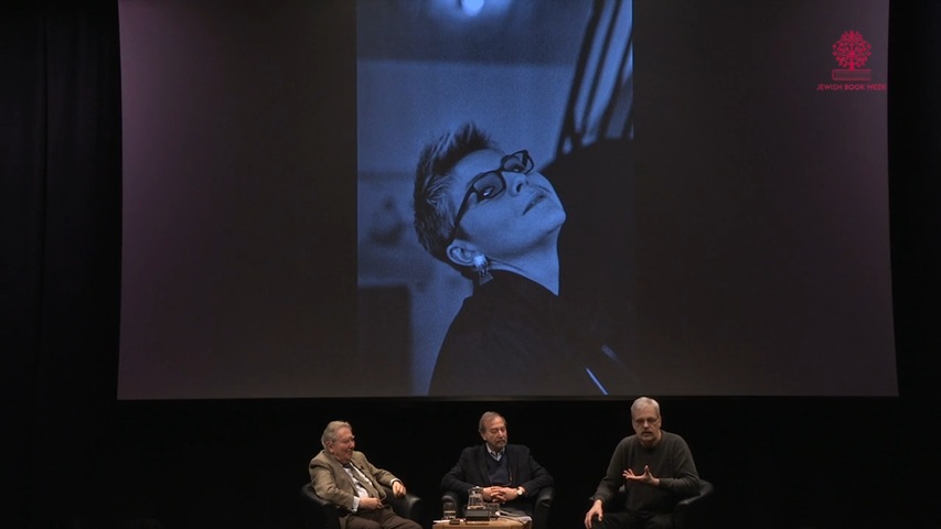 Three men sit on a dark stage facing the audience. Above and behind them, a blue-toned image of a woman with short hair and glasses is displayed.