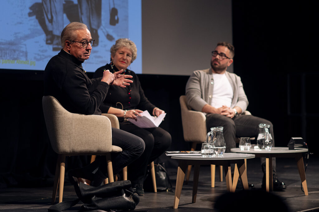 Two men and a woman sit on stage. One man is speaking and gesturing, while the other man and the woman watch him.