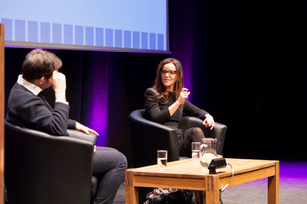 Two people sit on stage, one with their back to the camera. The other person, a white woman with dark hair and glasses, is speaking and gesturing.