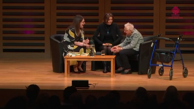 Three women sit around a table on a stage, leaning towards each other and smiling