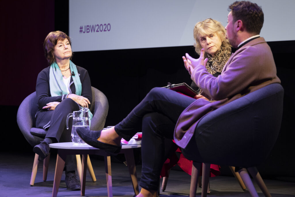 Two women and a man sit on a stage. The women look at the man who has his back to the camera and is gesturing with his hands