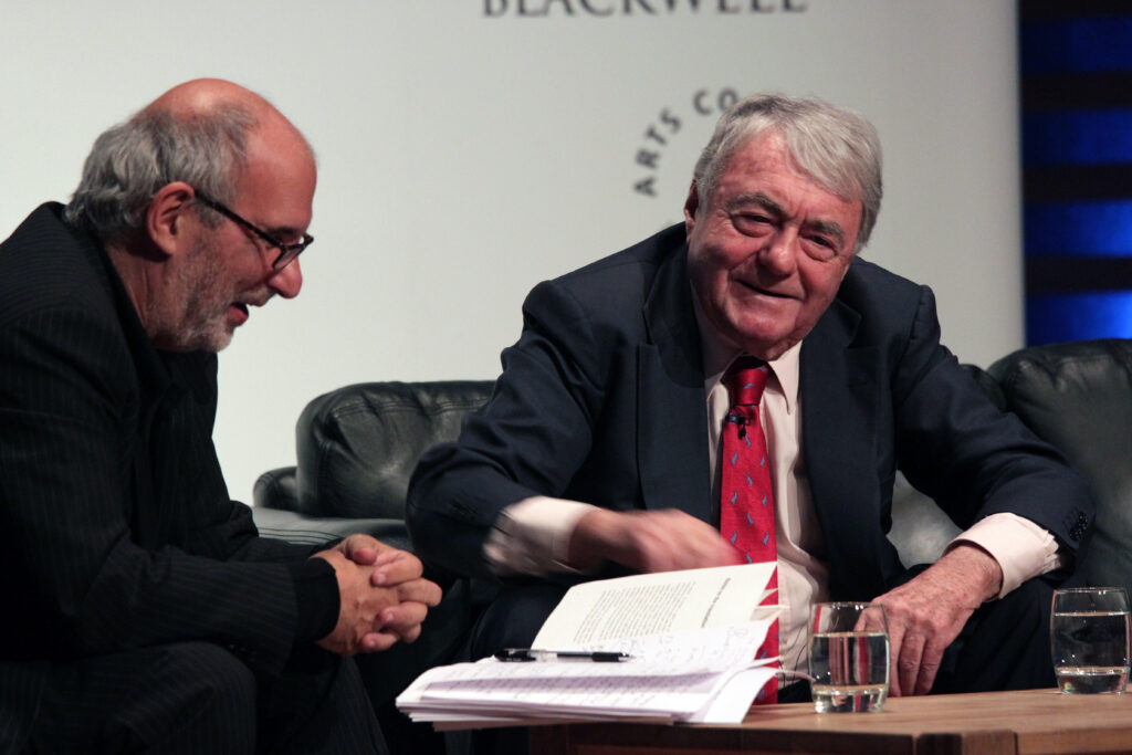 Two white men in suits sit at a table, a book open in front of them