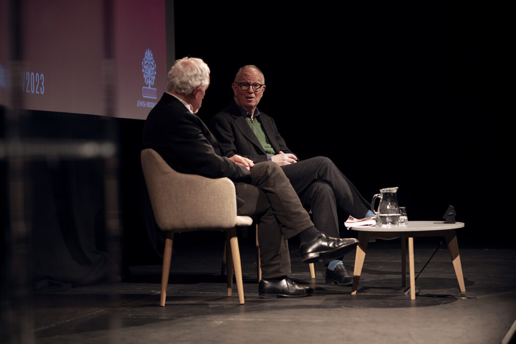 Two men sit on a dark stage. One has his back to the camera, the other is turned to face him.