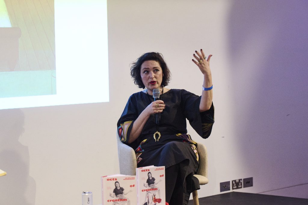 A seated woman wearing a black jumper and belt with a gold buckle speaks into a microphone while gesturing with her left hand held in the air.