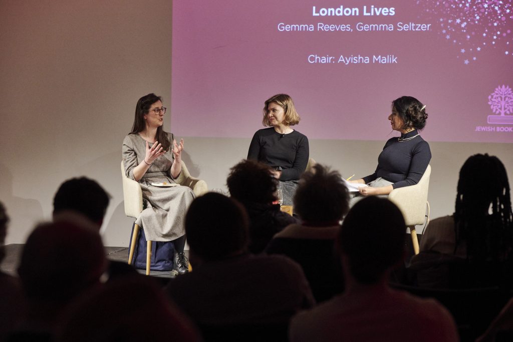 A group of three women seated on a stage. One speaks into a microphone while the other two listen
