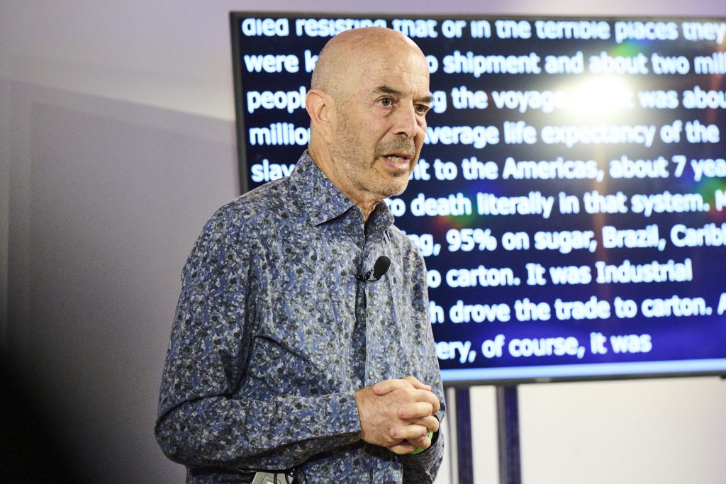 A white man speaks in front of an LCD screen.