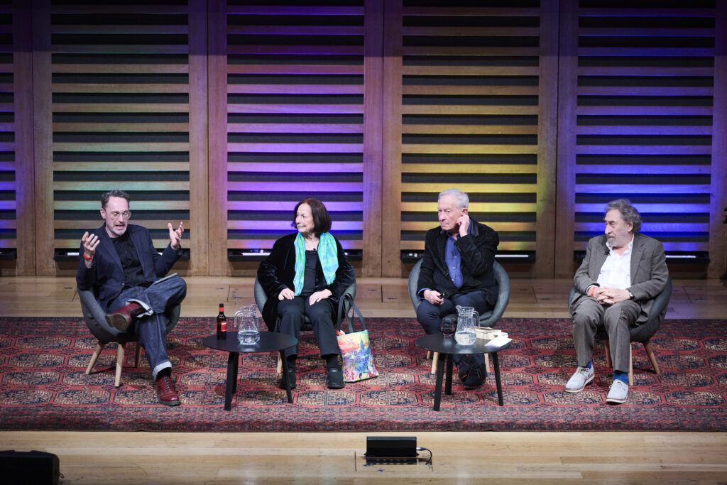 Three men and a woman sit on a stage in a line of chairs. They appear to be in lively discussion; the man on the far left is gesturing.