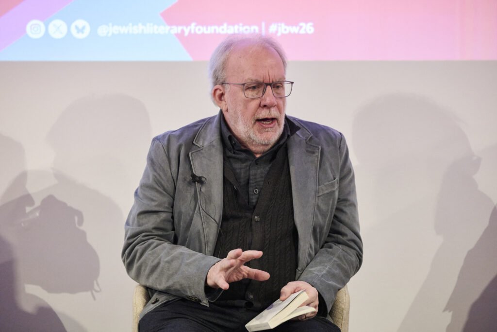 A white man is speaking, holding a book in his left hand and gesturing with the palm of his right hand down and fingers open. He is wearing a navy shirt and grey blazer, with thin-rimmed spectacles.