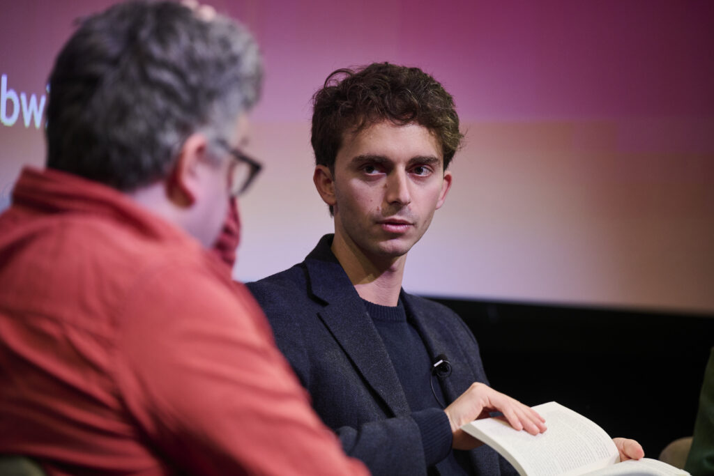 two men talking to each other, one man wearing a red shirt, other man wearing dark suit.
