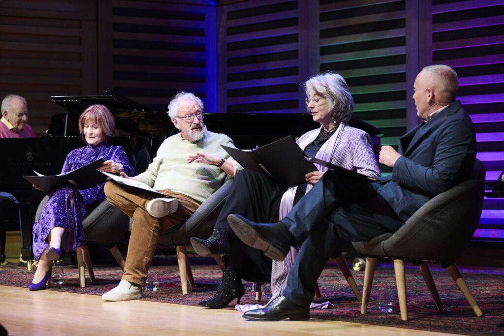 four people sitting with ring binders at live event, talking to each other on stage