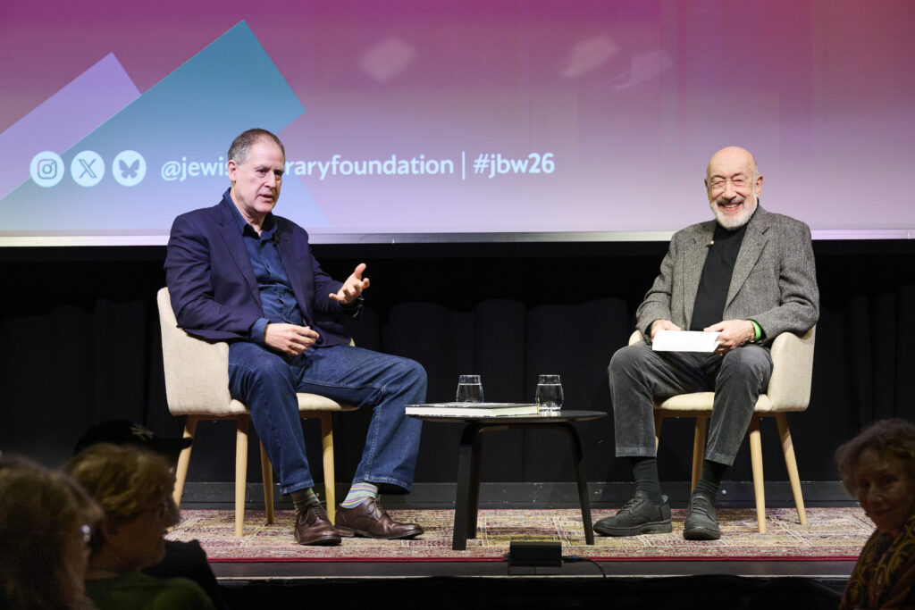 Two men sitting on chairs talking to each other in a live event, wearing suits