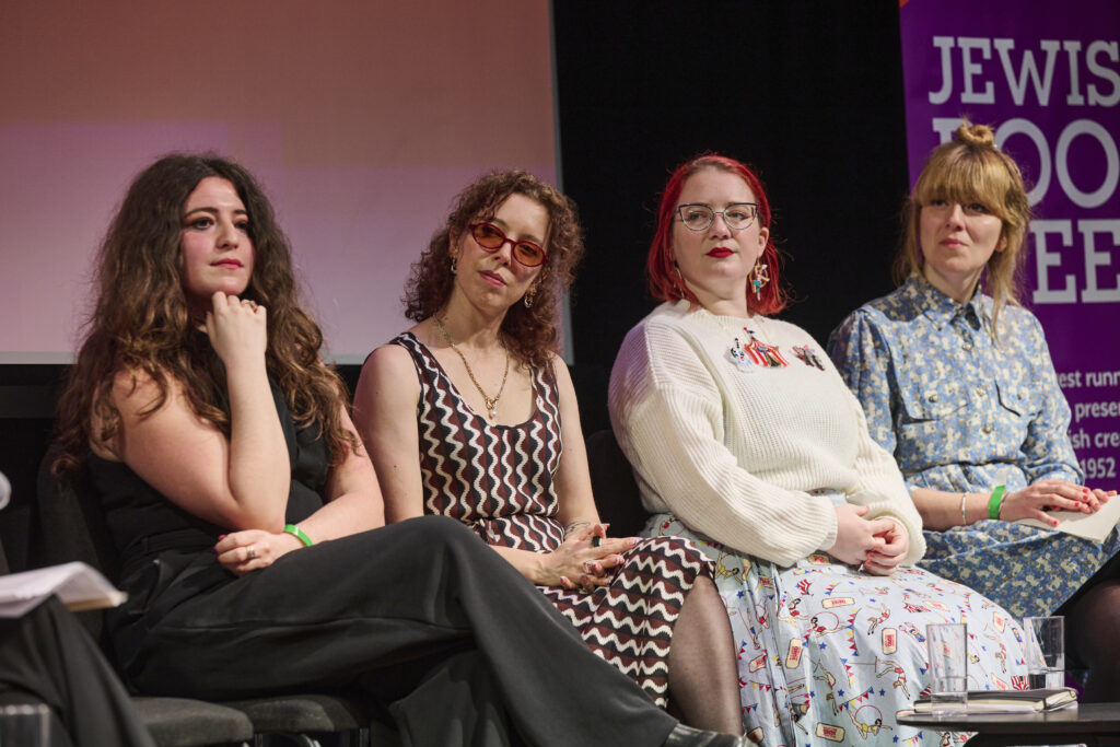 Four women speaking in live event, sitting on chairs with a pull up banner behind