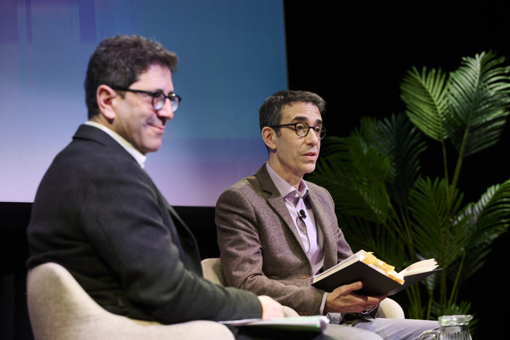two men sitting in chair talking with a book in their hand